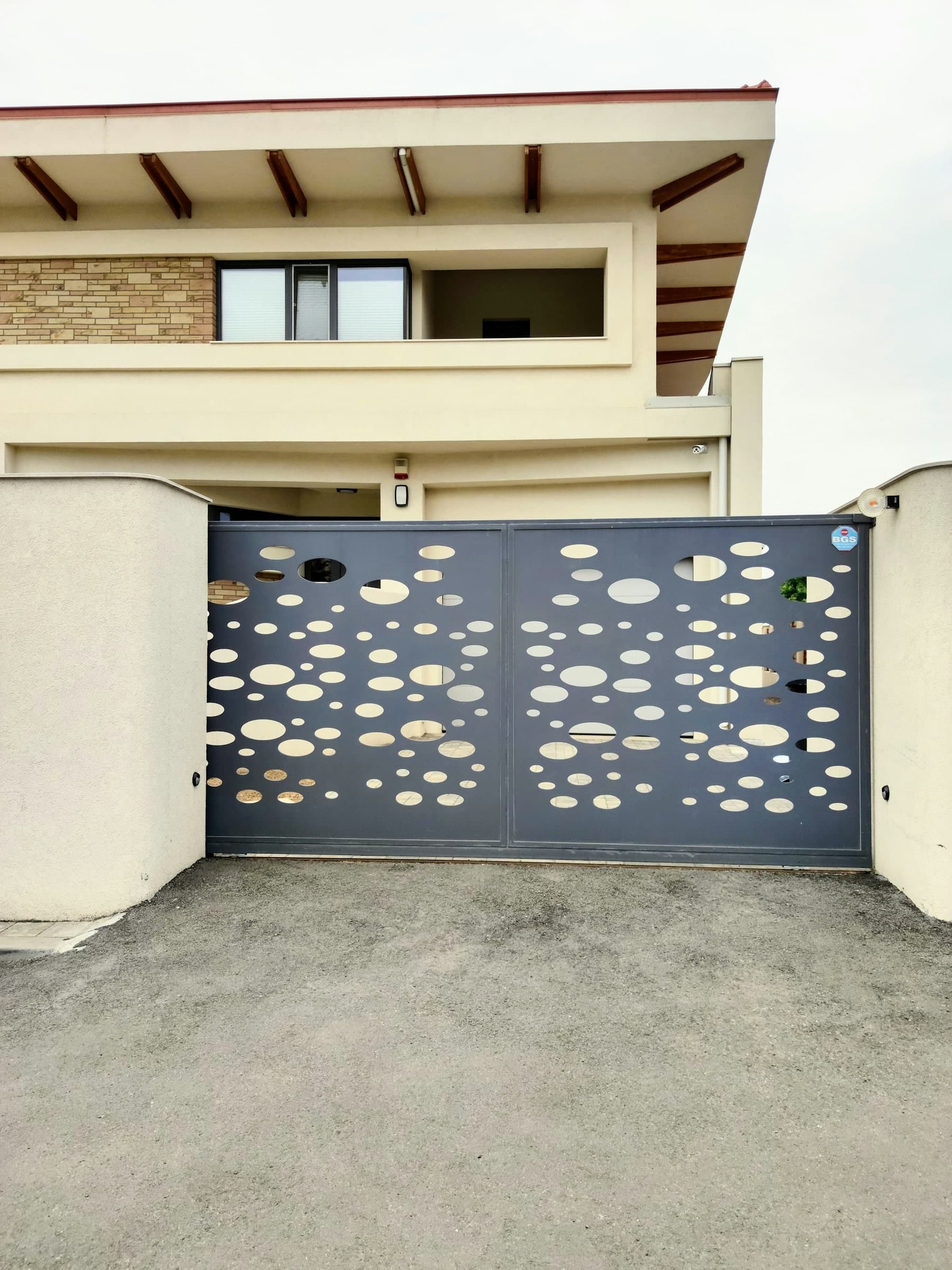 Grey perforated-panel modern gate across a driveway beneath a cantilevered house overhang.