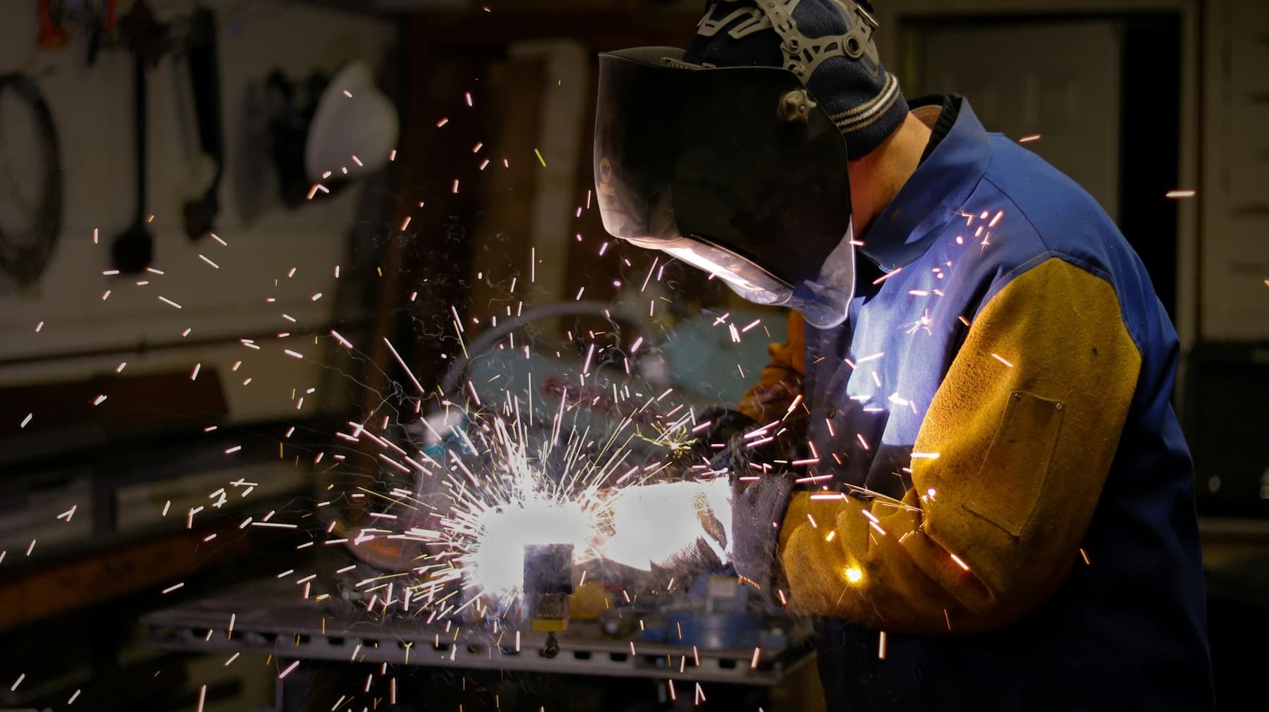 Welder in a hood laying a bead on steel with sparks flying across the bench