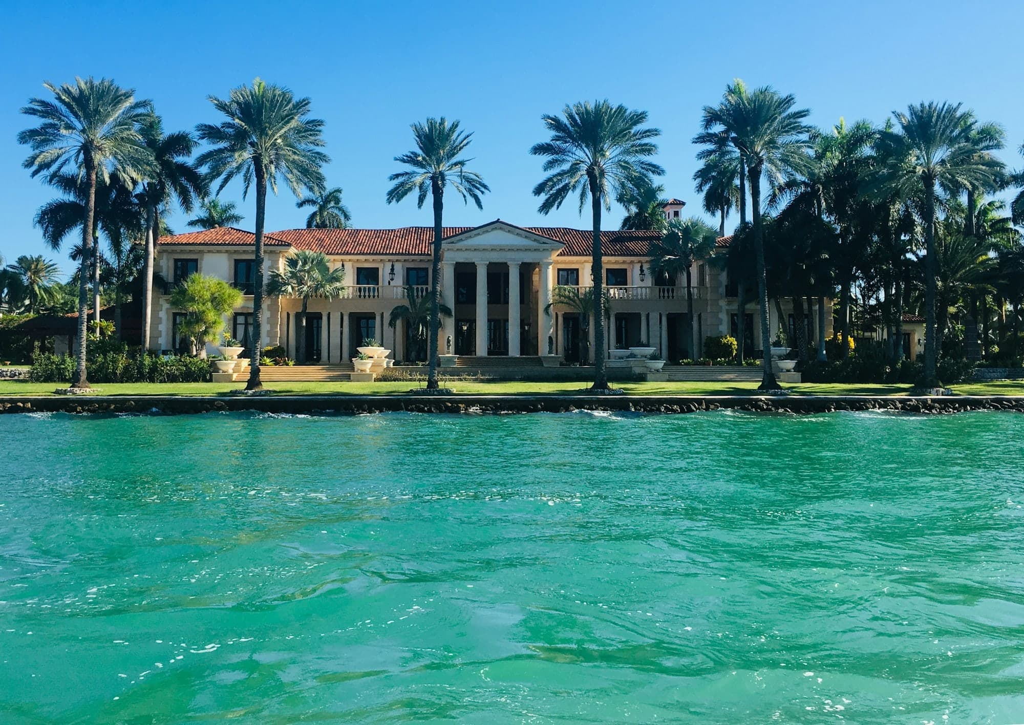 Waterfront estate seen from the water, its lawn lined with tall palms above a seawall.