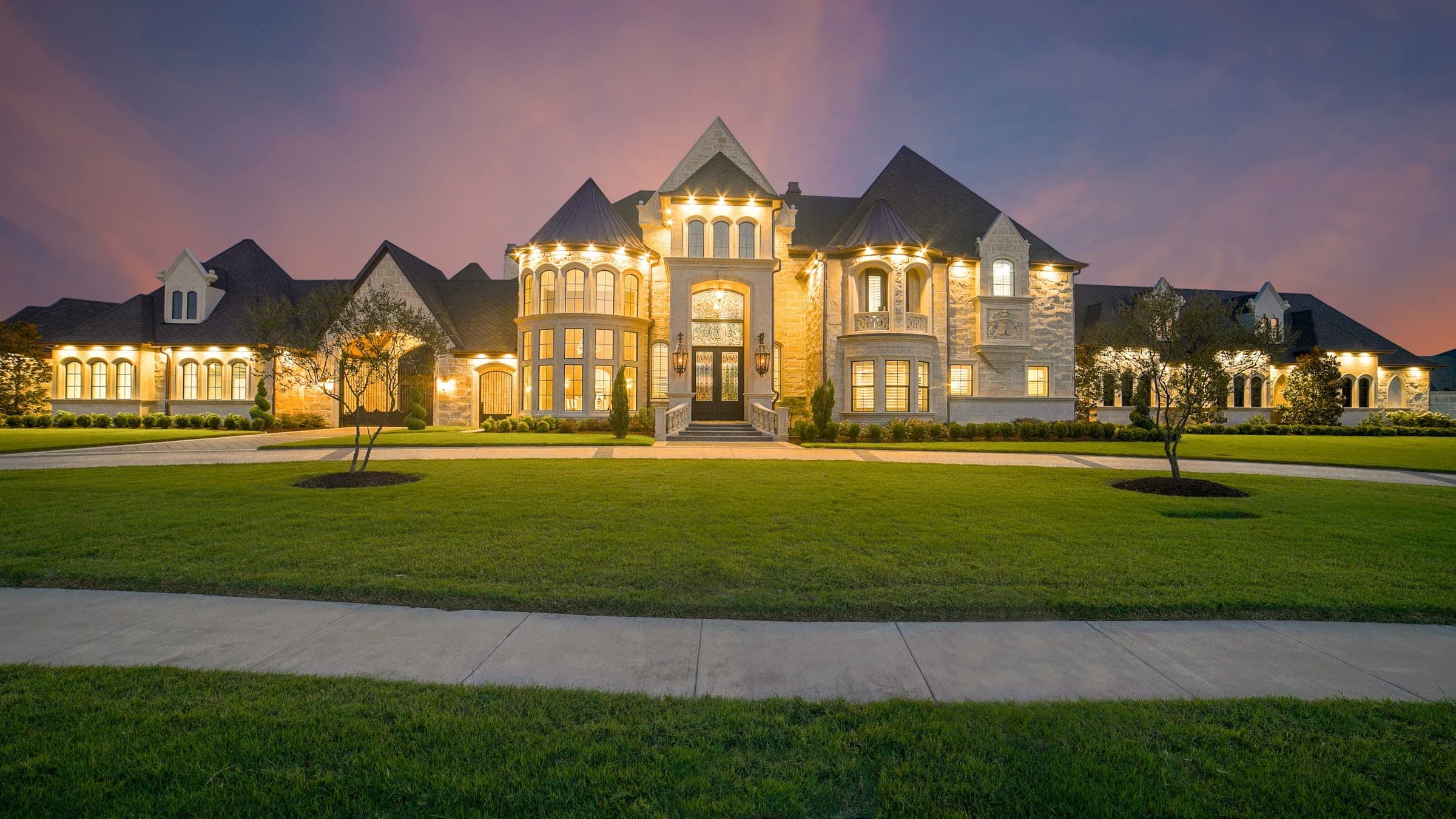 Large stone estate lit from within at dusk, seen across the front lawn