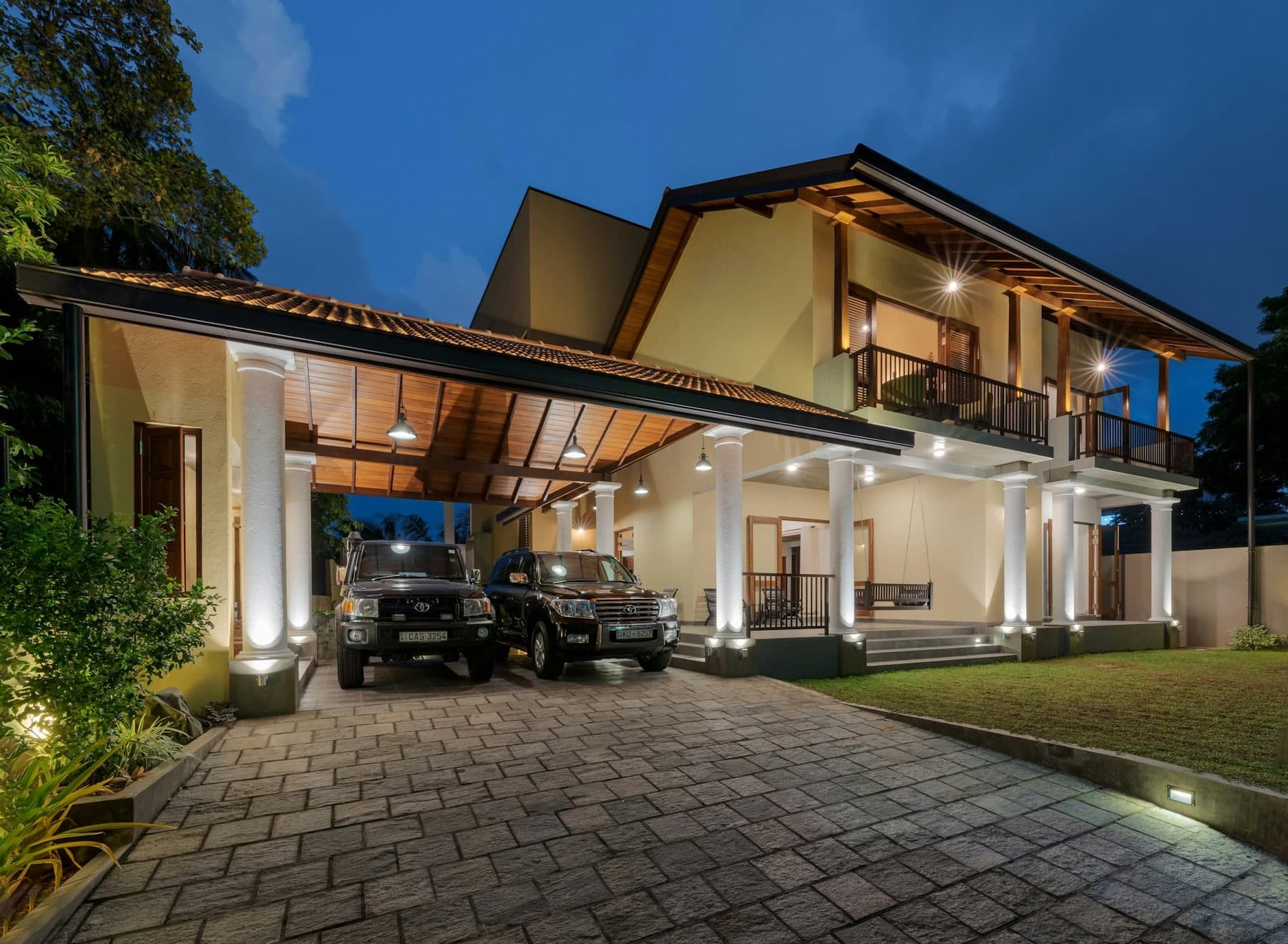 Lit modern house at dusk with vehicles parked under the entrance canopy